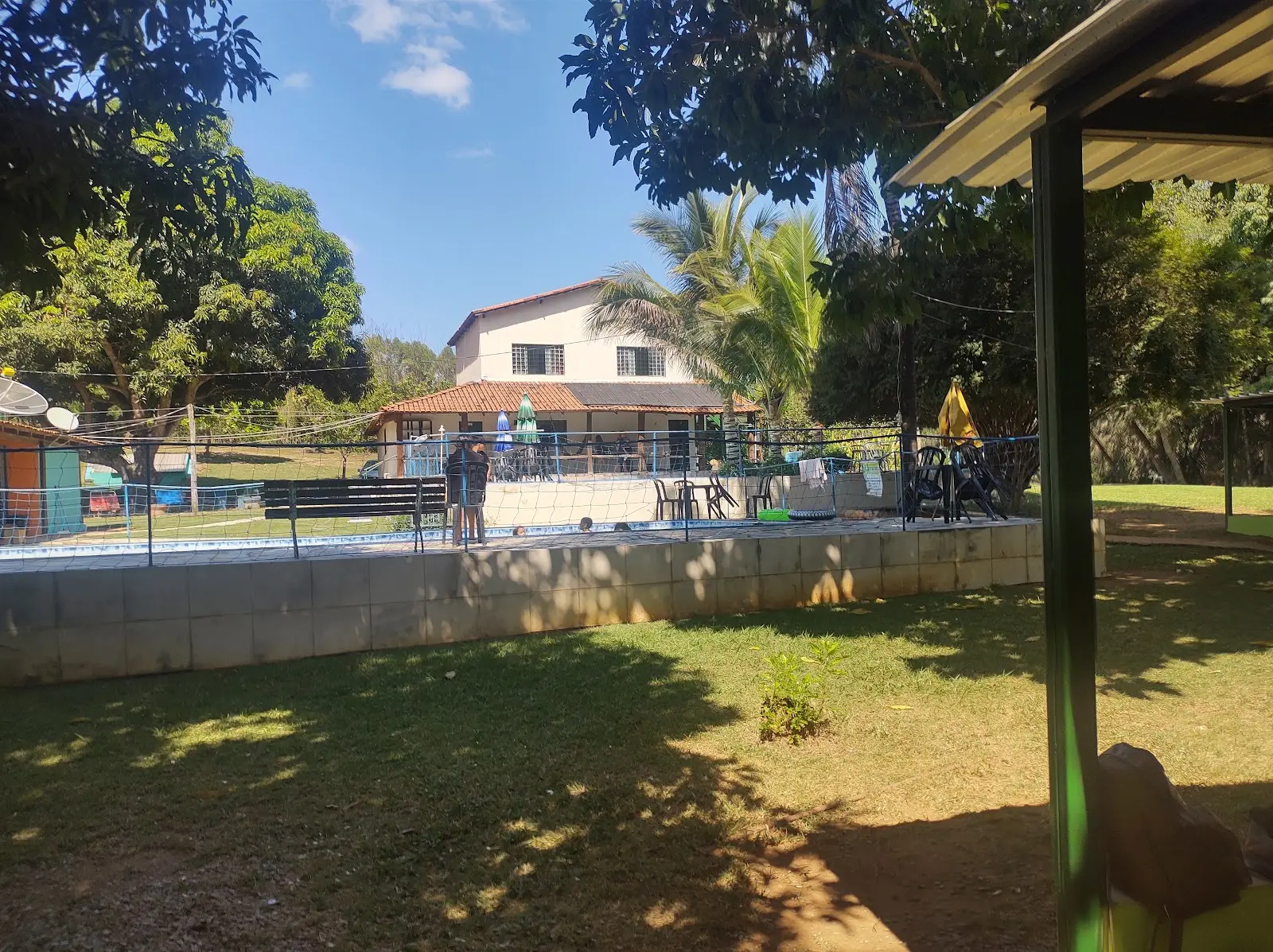 Piscina cercada por tela diante de uma casa de dois andares, com gramado, árvores e guarda-sóis