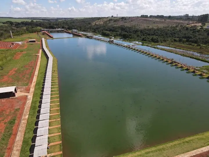 Vista aérea do lago comprido do Clube Pescar, com a fileira de cabanas brancas na margem e os decks de pesca do outro lado