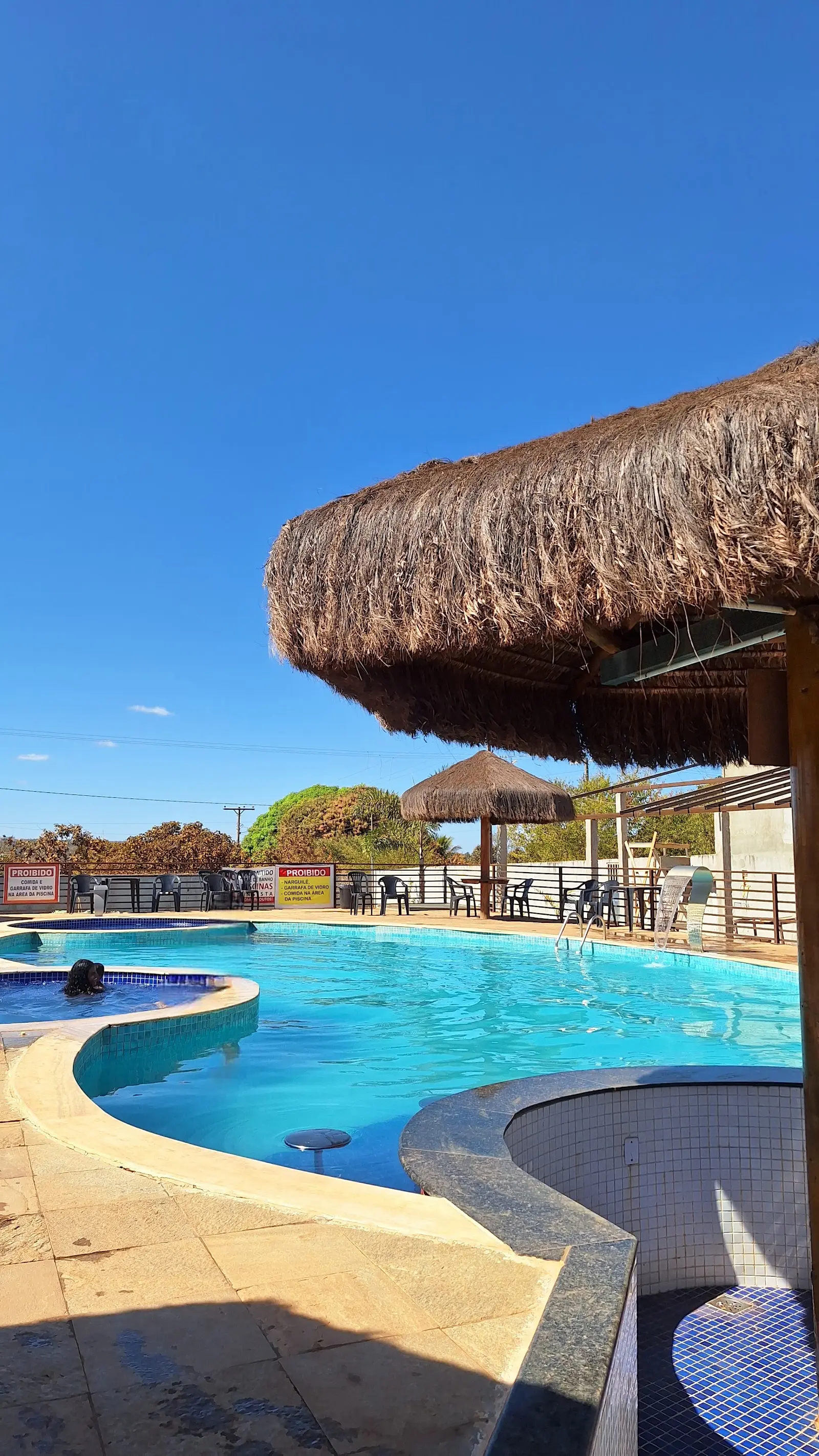 Piscina azul com quiosque de palha em primeiro plano e mesas ao fundo, sob céu limpo