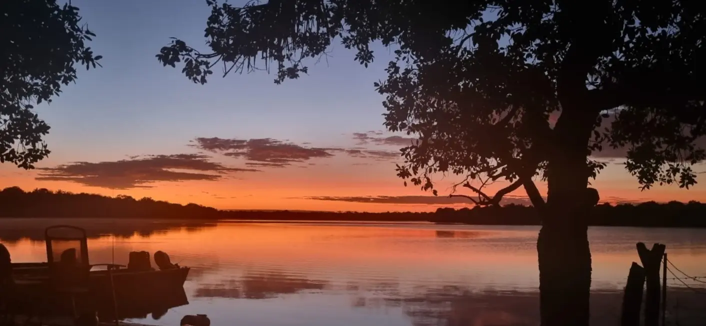 Pôr do sol sobre o lago da Fazenda Lago Azul, com barcos na margem e uma árvore em primeiro plano