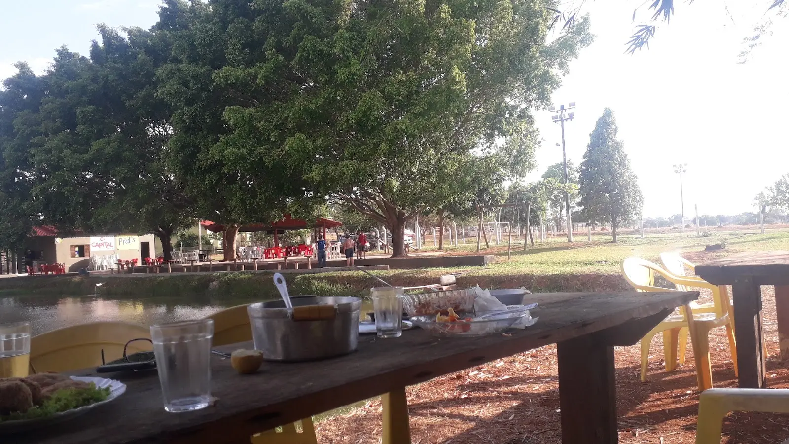 Mesa à beira do lago no Pesque Pague do Zé Marinho, com árvores grandes e mesas vermelhas na outra margem