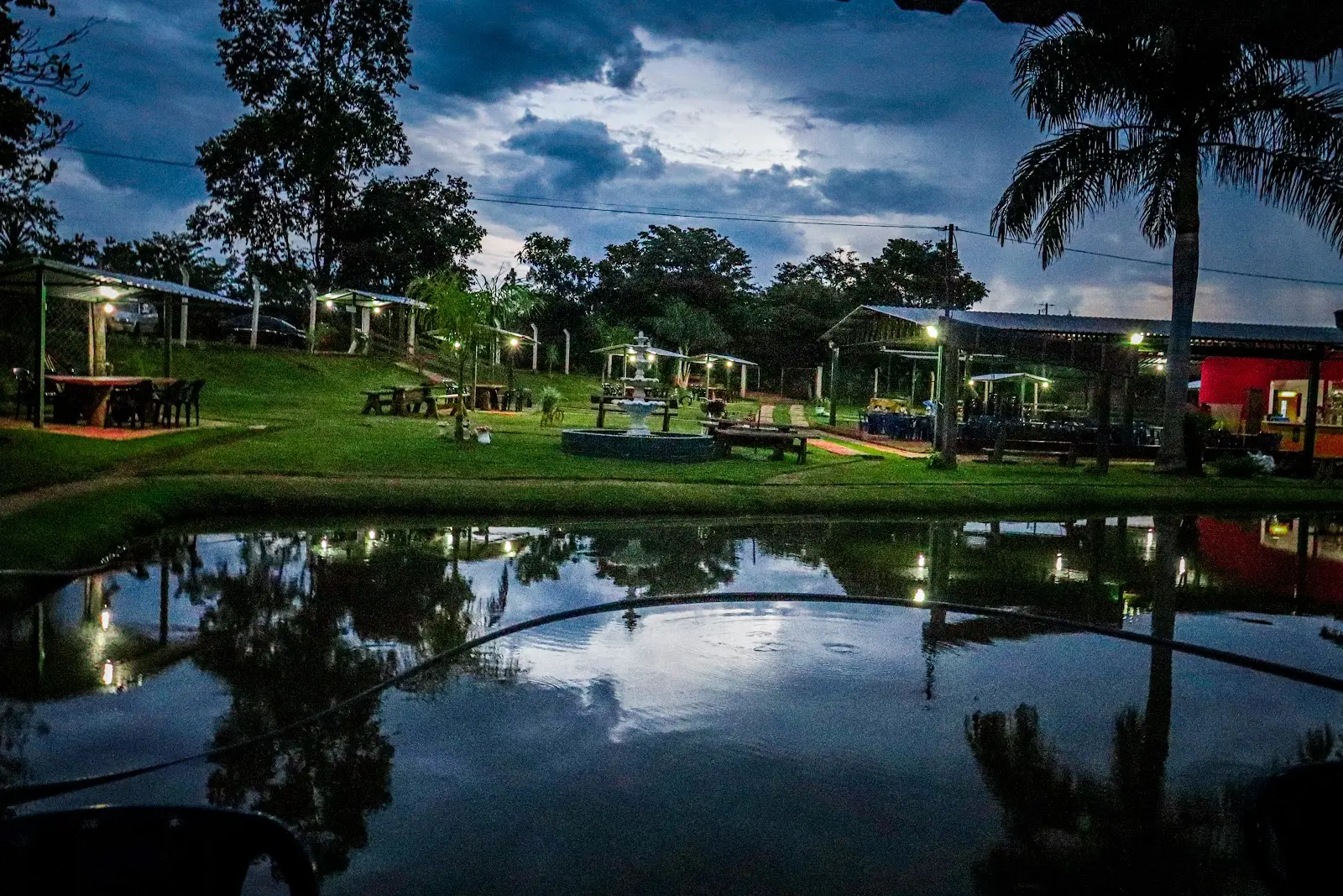 Lago do Pesque e Pague do Gordinho ao anoitecer, com quiosques iluminados e uma fonte no gramado