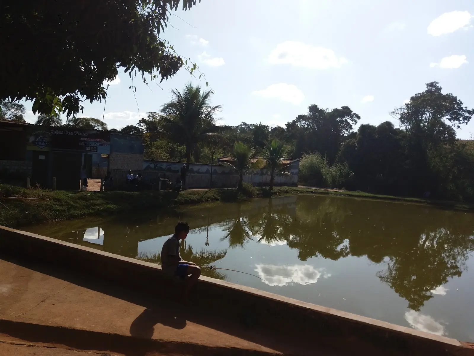 Menino pesca sentado na mureta de um lago cercado de árvores no Pesque e Pague El Shadai