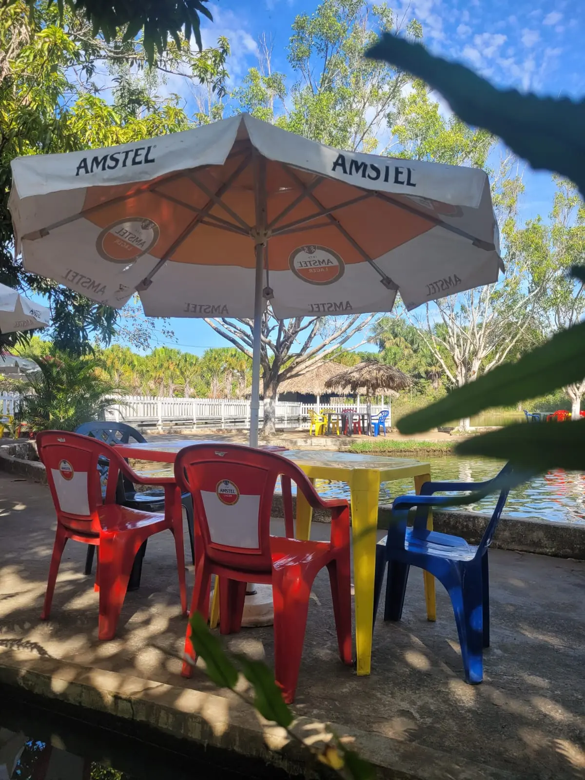 Mesa com guarda-sol à beira do tanque no Pesque Pague Água Doce, com quiosques de palha ao fundo