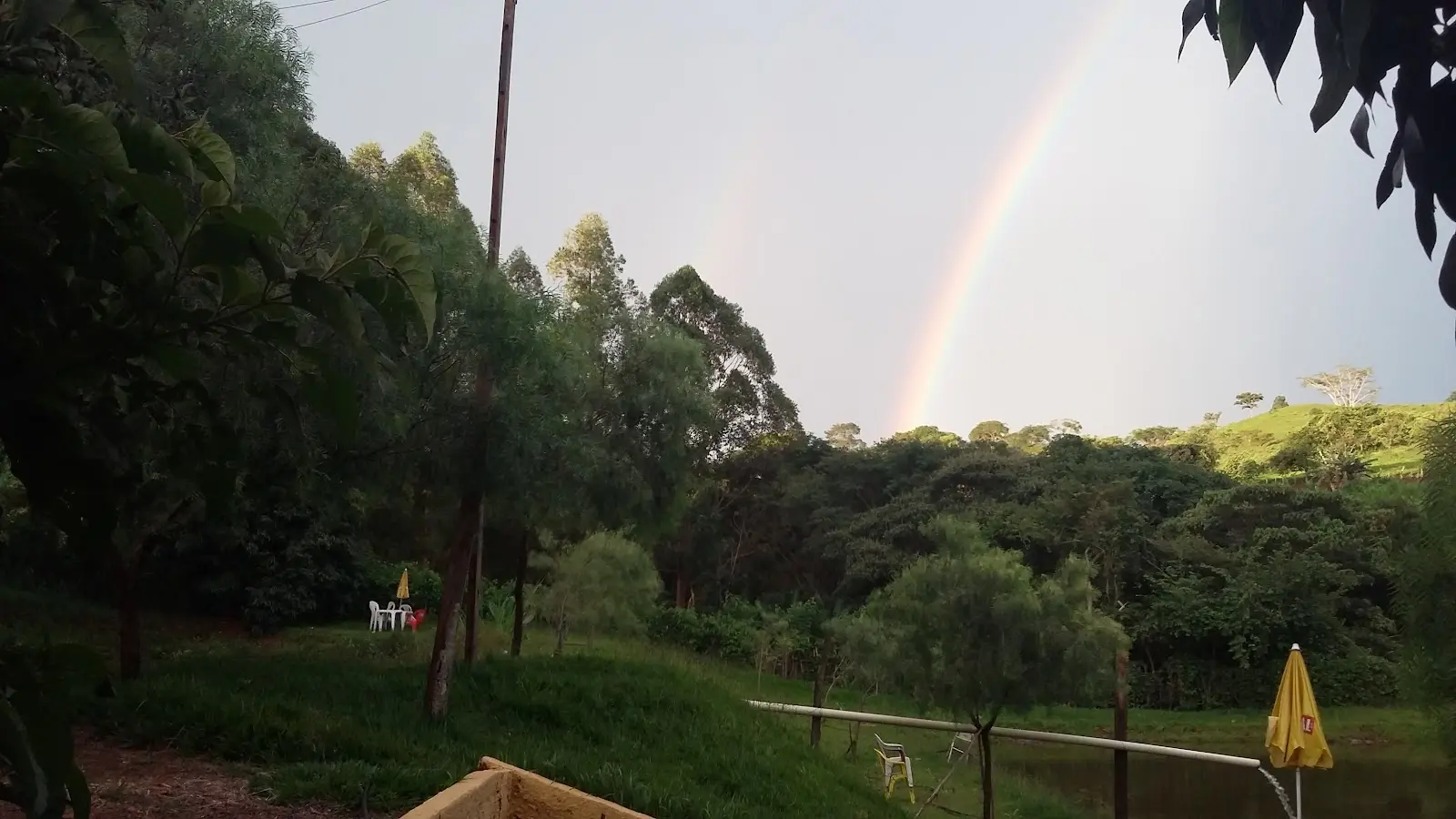 Arco-íris sobre a mata do Pesque Pague Araújo, com um pedaço do lago e um guarda-sol amarelo