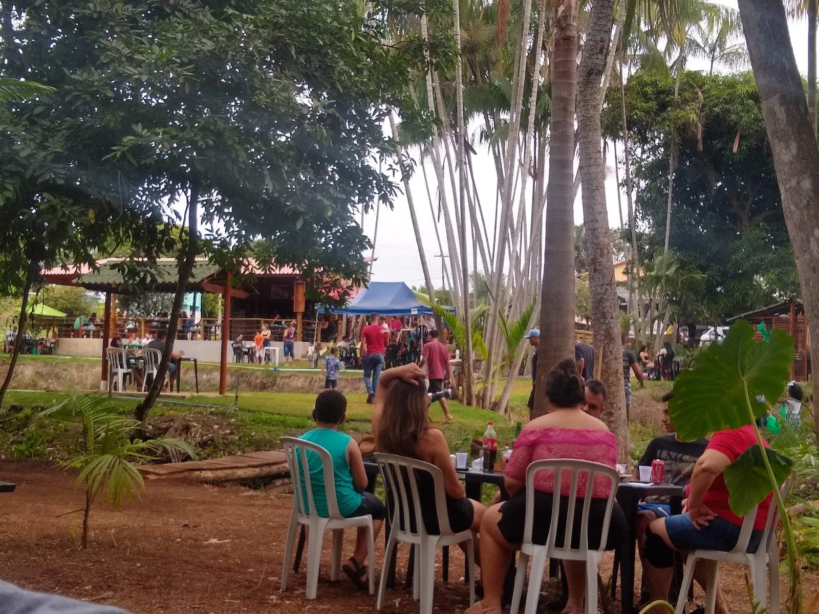 Clientes em mesas plásticas sob as árvores, com uma tenda e um quiosque de madeira ao fundo