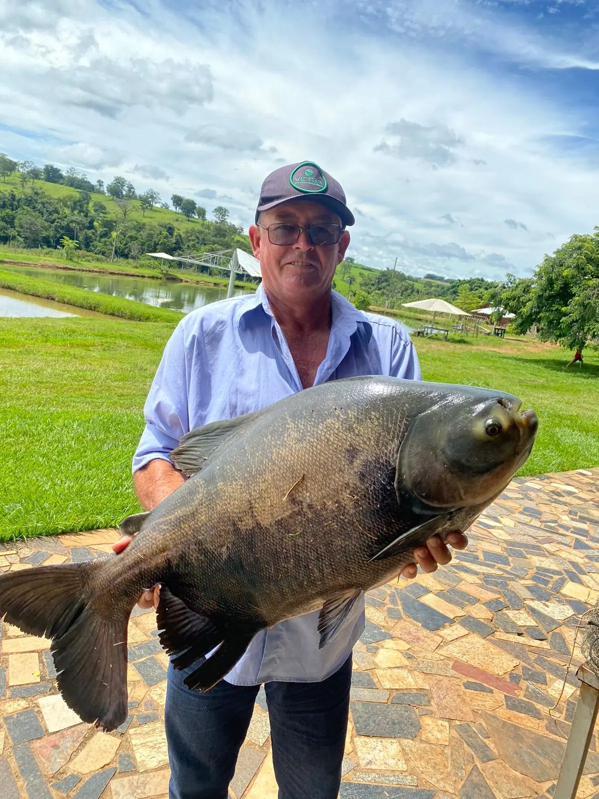 Pescador segurando um peixe redondo grande, com os tanques e guarda-sóis do Pesque Pague do Dirceu ao fundo