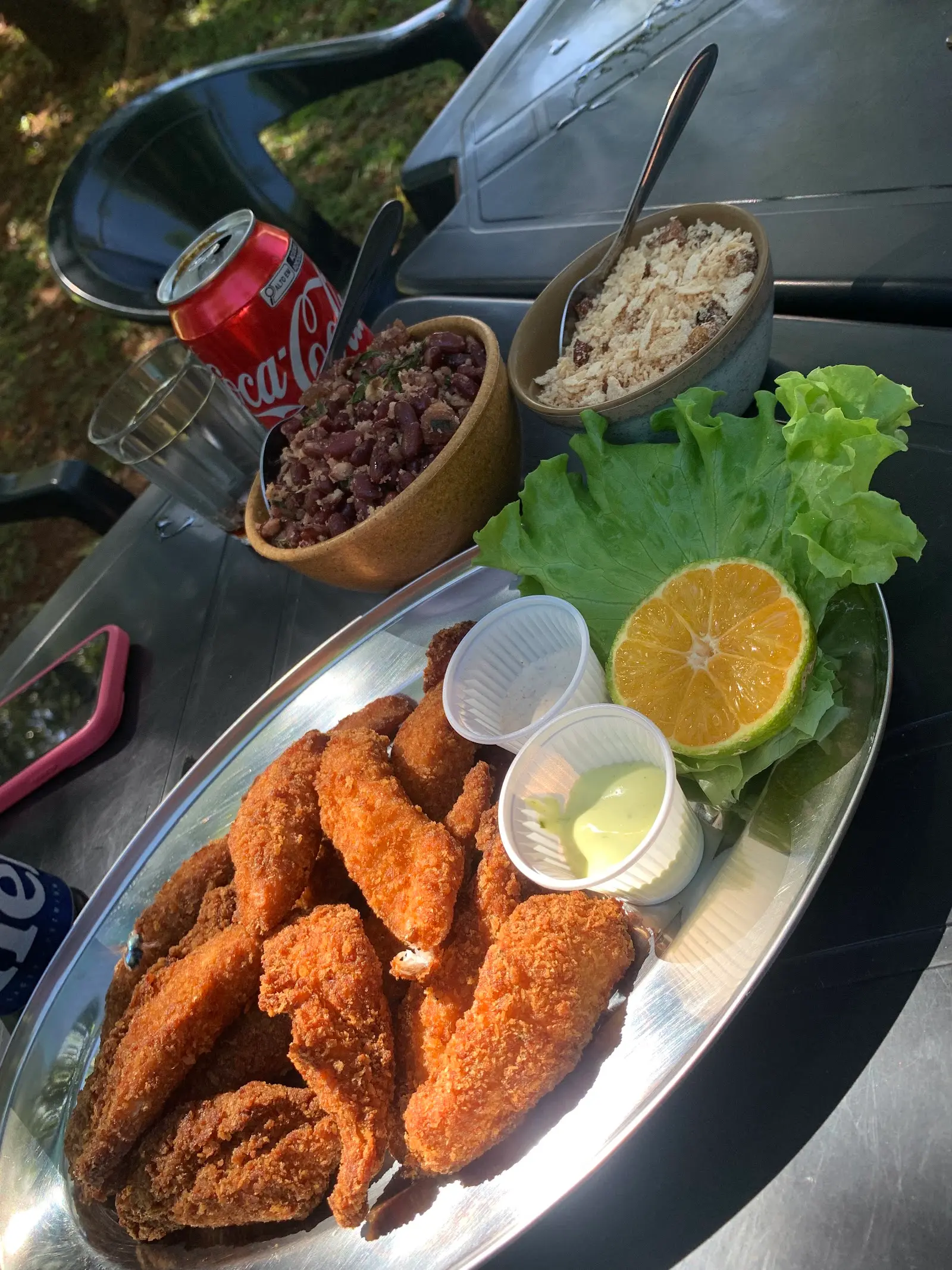 Travessa de peixe empanado com limão, feijão tropeiro e farofa numa mesa ao ar livre