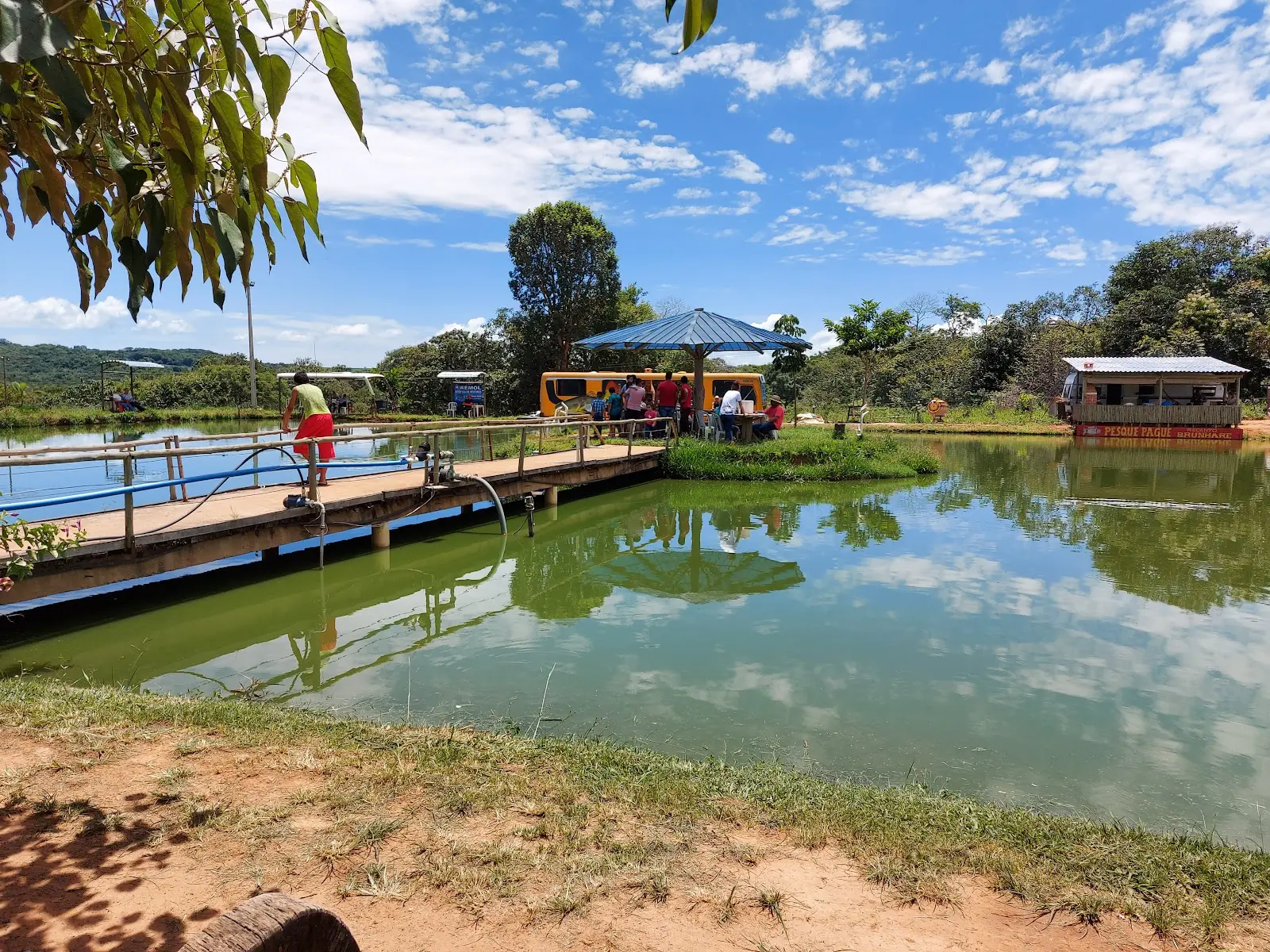 Lago do Quintal do Vidal com píer de madeira, guarda-sol e um ônibus adaptado ao fundo