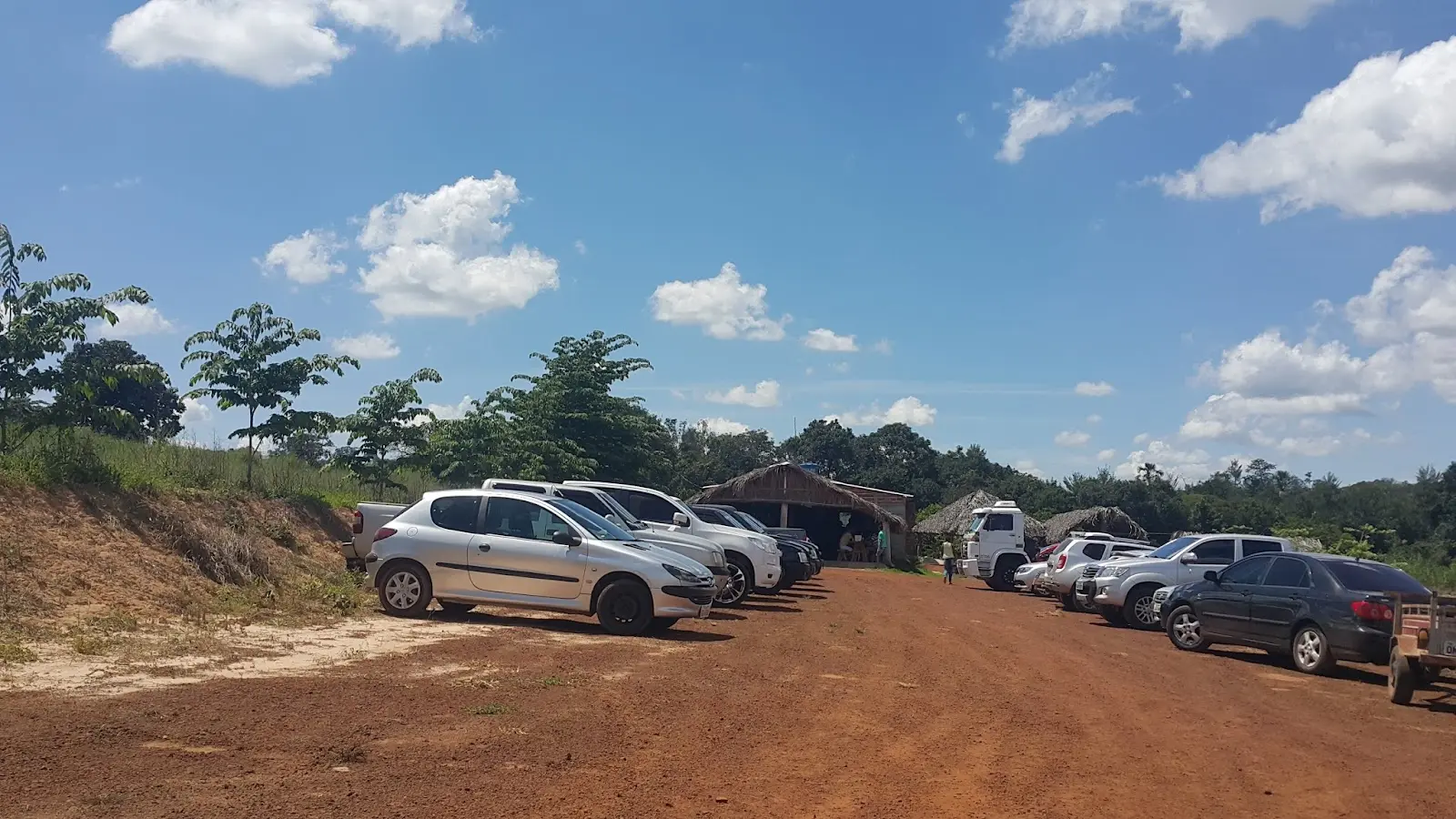 Estacionamento de terra cheio de carros e quiosques cobertos de palha no Pesque Pague Xavier