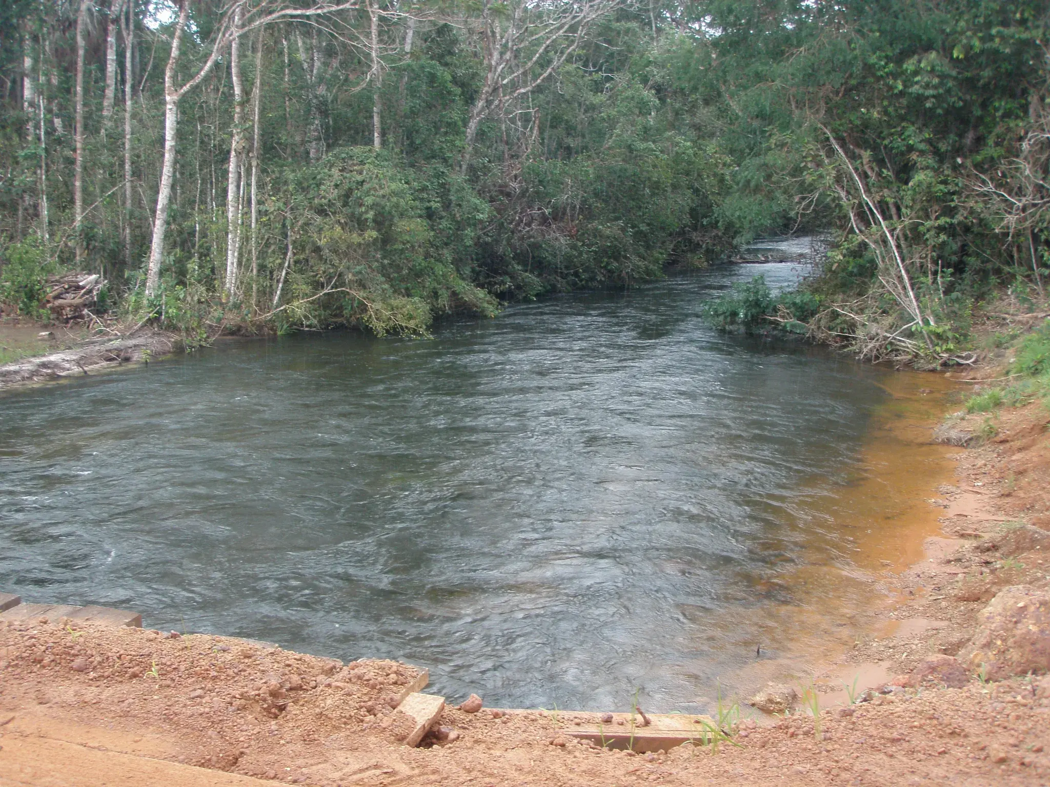 Rio de correnteza entre a mata, visto da margem de terra vermelha junto à estrada