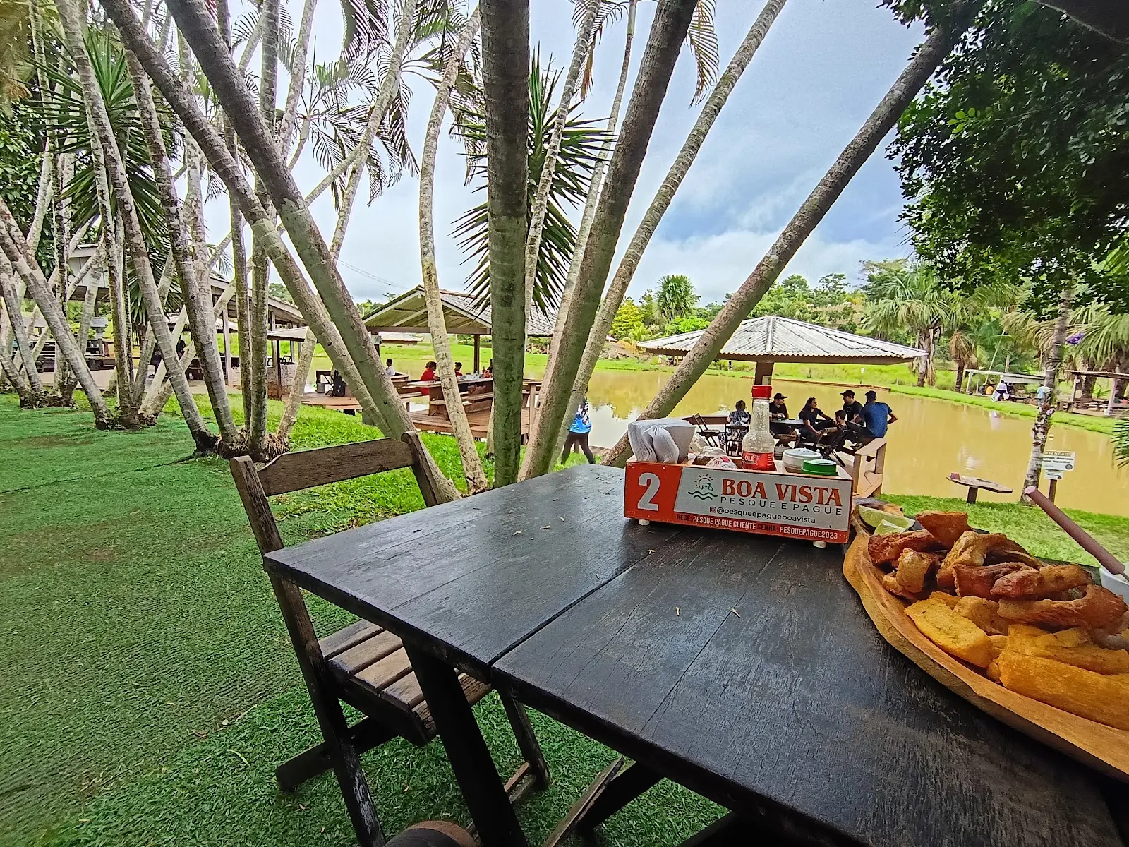 Mesa com porção de peixe frito e mandioca, com o lago e quiosques cheios de gente ao fundo, no Pesque e Pague Boa Vista em Cacoal